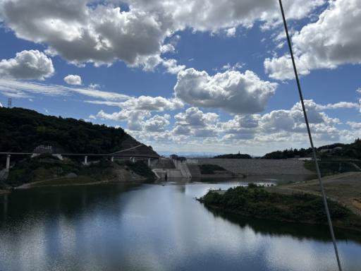 A serene landscape featuring a large body of water reflecting the sky. In the background, a dam stretches horizontally across the image, flanked by green hills. Fluffy clouds float across a bright blue sky, casting shadows on the land and water below. A cable is visible in the foreground.