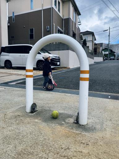 A young child wearing a cap and dark jacket stands near a white U-shaped barrier with orange stripes, holding a colorful mesh bag. A tennis ball lies on the pavement in front of the barrier. In the background, there is a white van parked beside a residential building on a cloudy day.