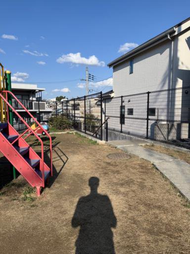 A playground with a red and blue slide is in the left foreground. My shadow extends onto the dry grass. A clear blue sky with a few clouds is visible above, with a utility pole and wires in the distance.
